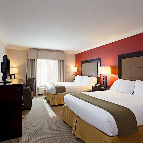 This image shows a hotel room with two large beds, a TV on a dresser, a chair by the window, and a red accent wall behind the beds.