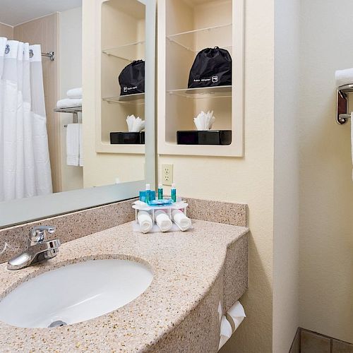 A clean bathroom with a sink, mirror, toiletries, towels, a toilet, and a shower with a curtain is shown.