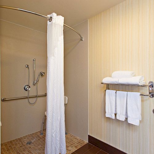 An accessible bathroom with a shower area, equipped with grab bars, a handheld shower head, a shower curtain, and a towel rack holding towels.