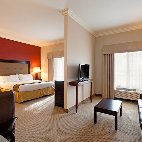 A modern hotel room with a king-size bed, a desk chair, a TV on a stand, a coffee table, and a sofa, featuring warm lighting and neutral decor.