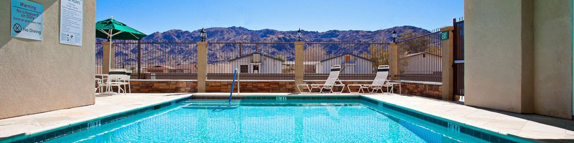 An outdoor swimming pool under a shaded structure with mountains in the background and lounge chairs beside the pool.