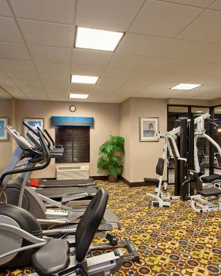 The image shows a well-lit gym with exercise equipment, including ellipticals, stationary bikes, and weight machines, and a TV mounted on the wall.