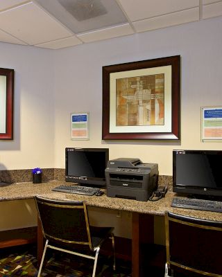 The image shows a hotel business center with two desktop computers, printers, framed artwork on the walls, and chairs for guests to use.