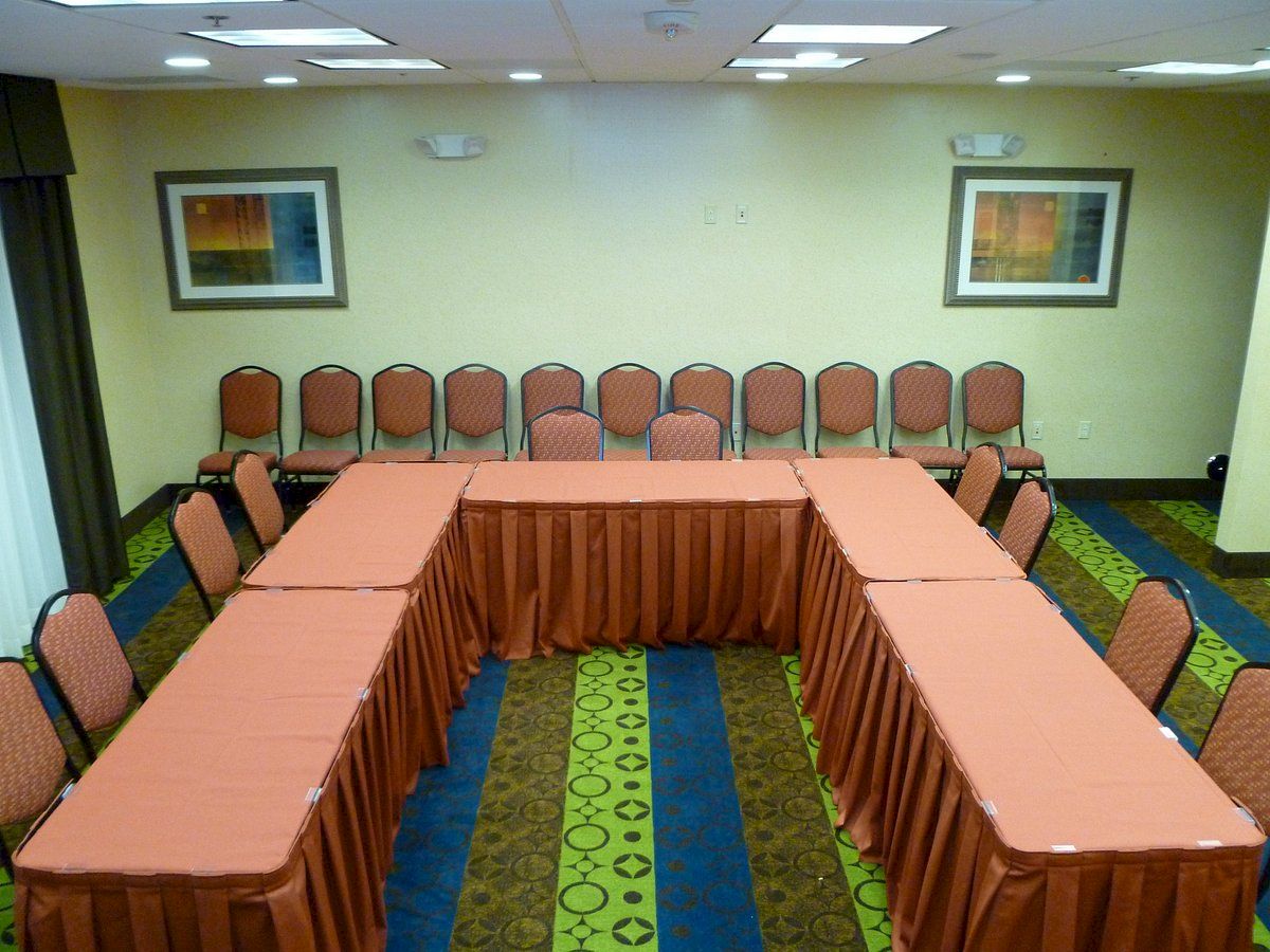 A conference room setup with U-shaped tables covered in orange cloths, surrounded by orange chairs, with artwork on the walls.