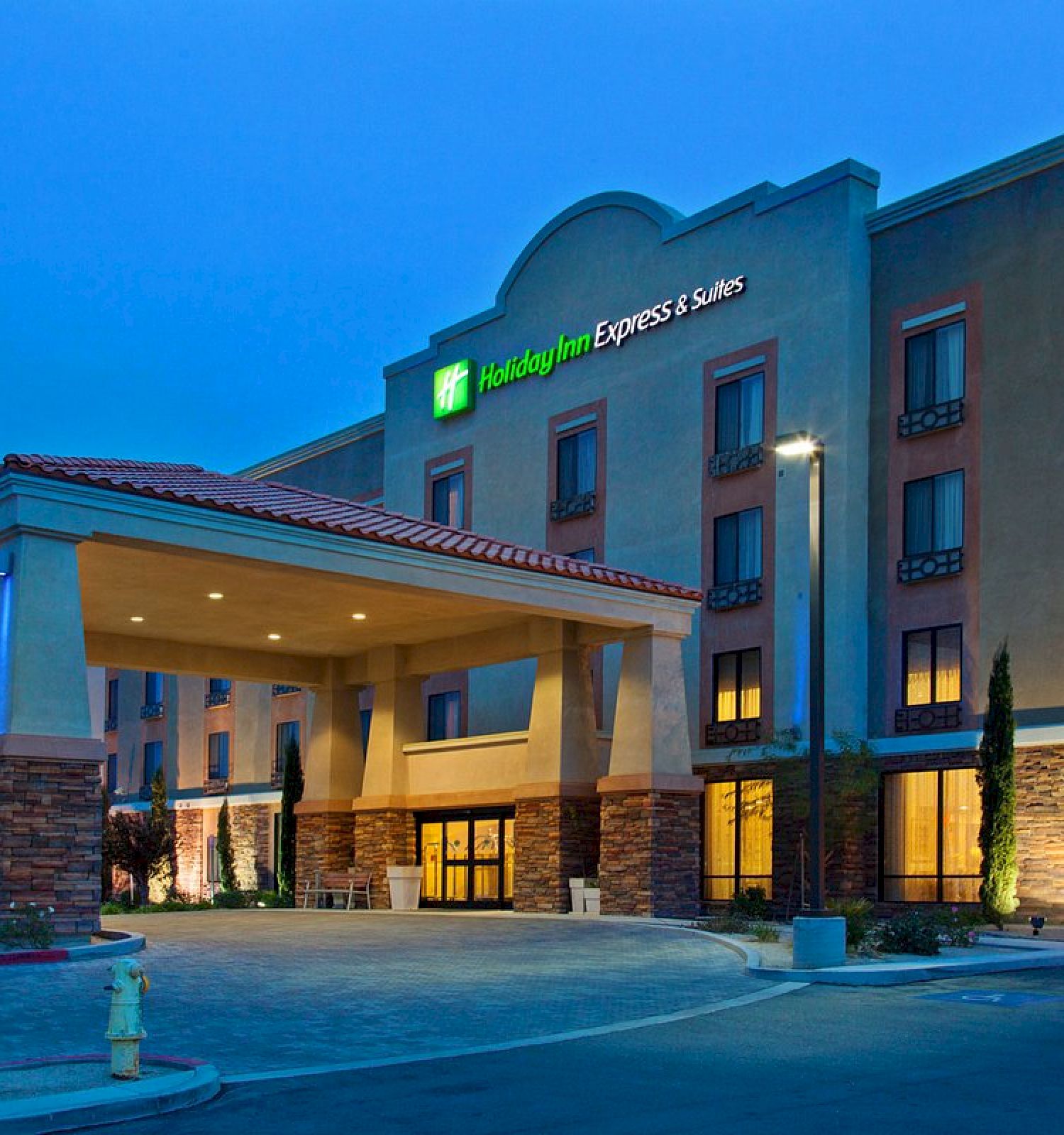 This image shows the exterior of a Holiday Inn Express & Suites hotel at dusk with lit-up signage, an entrance canopy, and surrounding landscaping.