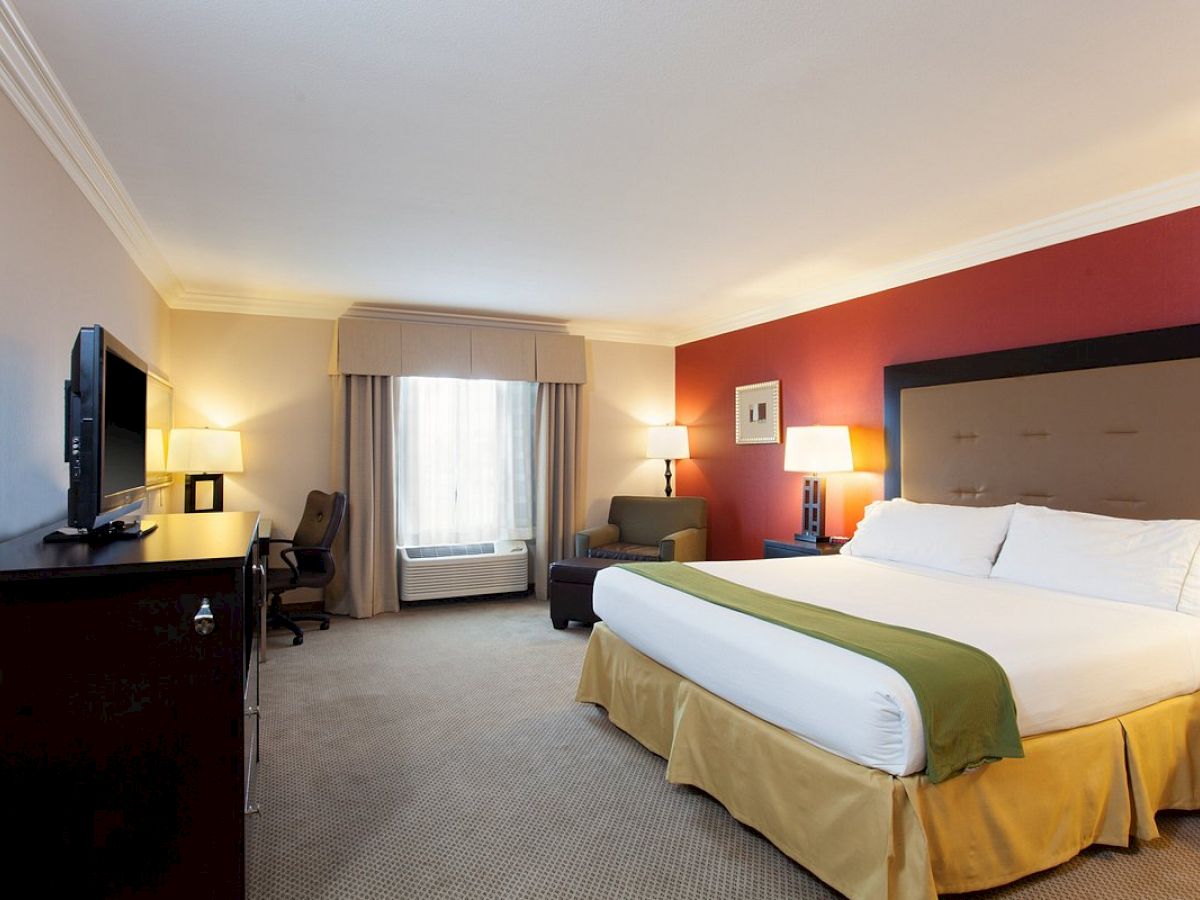 A hotel room with a large bed, TV, and an armchair, featuring modern décor and a red accent wall, warmly lit by table lamps.