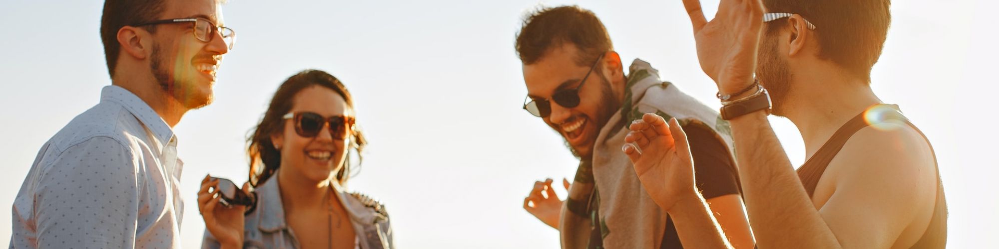 Four people are outdoors, laughing and conversing in a sunny setting. They appear to be enjoying each other's company and the pleasant weather.