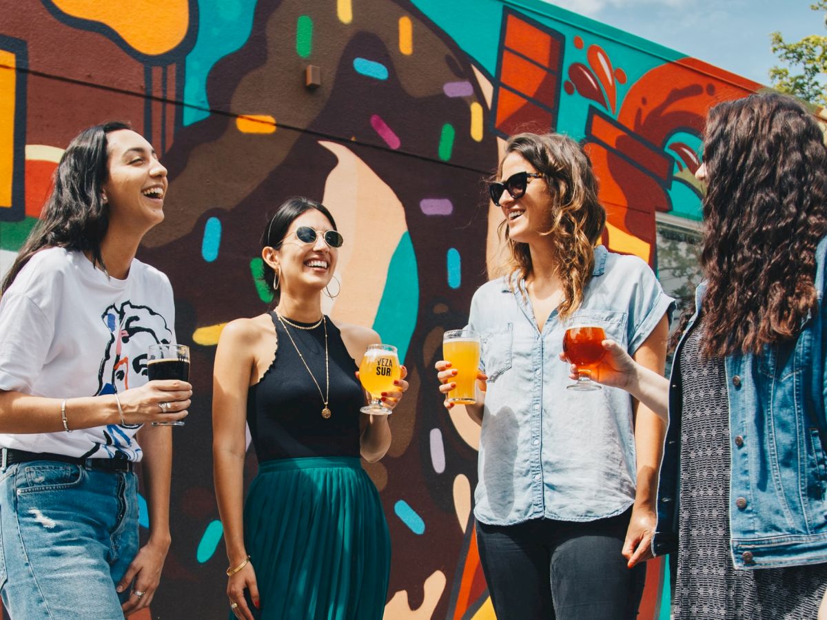 Four people are standing in front of a vibrant mural, enjoying drinks and laughing together, creating a joyful and colorful scene.