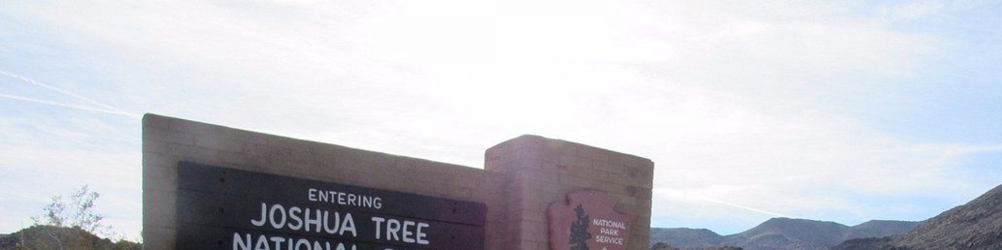 The image shows the entrance sign for Joshua Tree National Park, with a scenic mountain landscape and desert vegetation in the background.