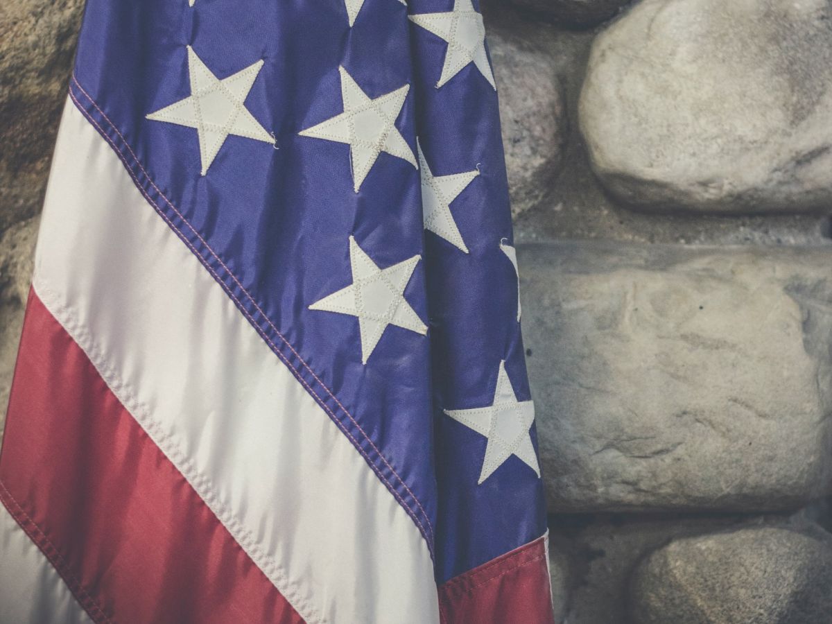 This image shows a close-up of an American flag with a stone wall background, featuring stars and stripes in red, white, and blue.