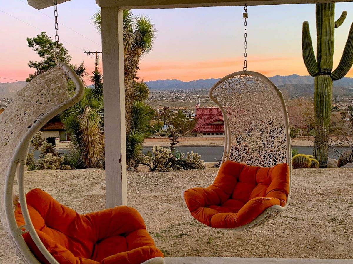 The image shows two hanging chairs with orange cushions on a patio, overlooking a desert landscape with a cactus and mountains in the distance.