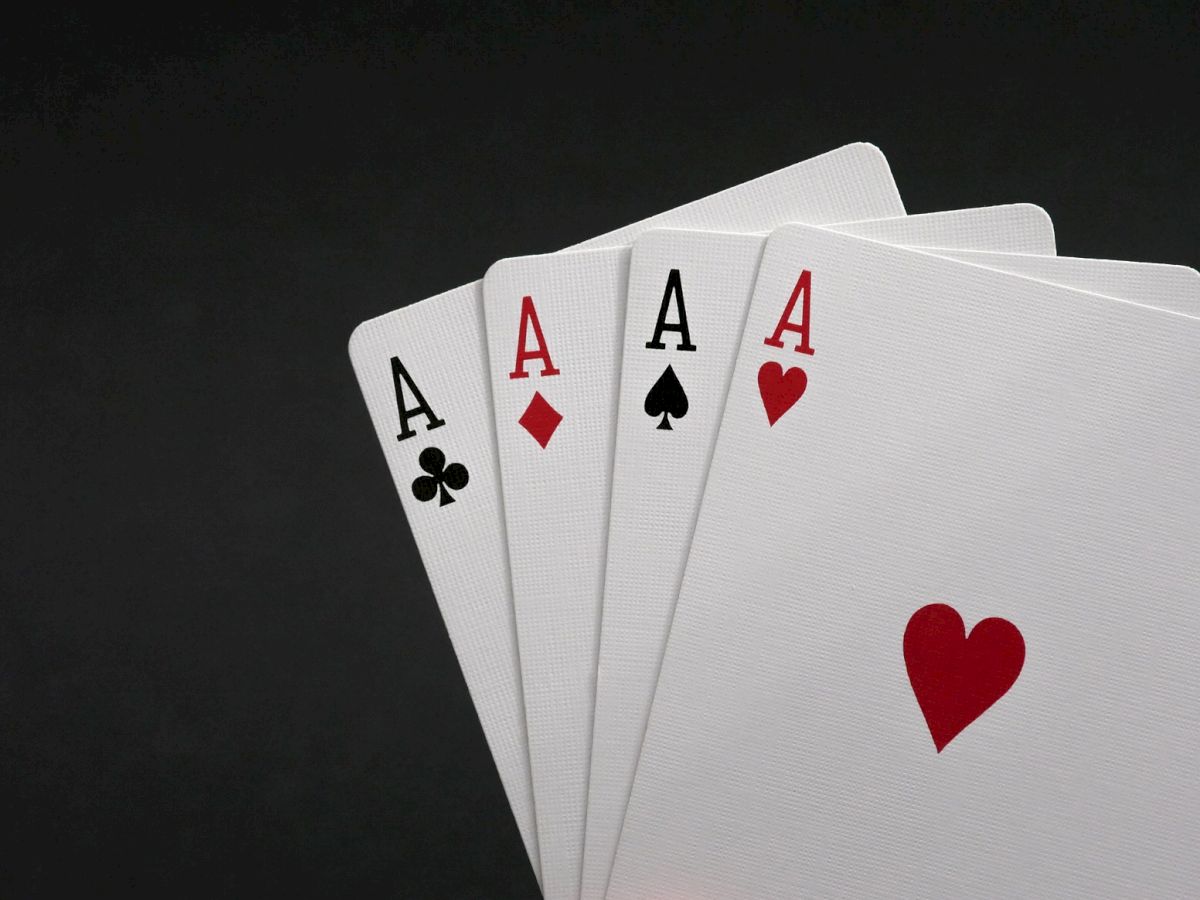 The image shows four playing cards arranged in a fan: Ace of Clubs, Ace of Diamonds, Ace of Spades, and Ace of Hearts, on a black surface.