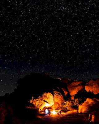 A starry night sky over a rocky landscape with a campfire illuminating a small area and possibly a person near the fire, creating a cozy scene.