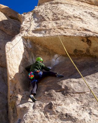 A person wearing climbing gear and a green helmet is rock climbing on a steep cliff, secured by a rope against a clear blue sky.