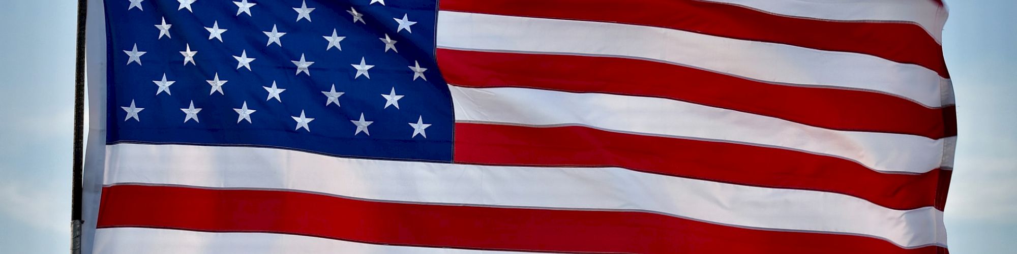 The image shows the flag of the United States of America flying against a blue sky, featuring stars and stripes.