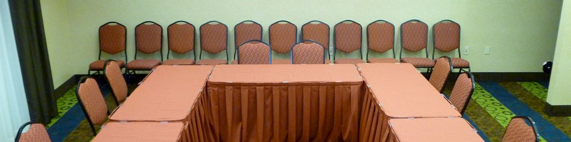 A conference room set up with a U-shaped table arrangement covered in red tablecloths, surrounded by red chairs. Two framed pictures hang on the wall.