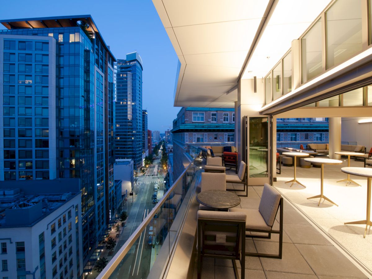 An upscale cityscape at dusk with a view from a modern balcony featuring outdoor seating and a glass railing, overlooking lit buildings and streets.