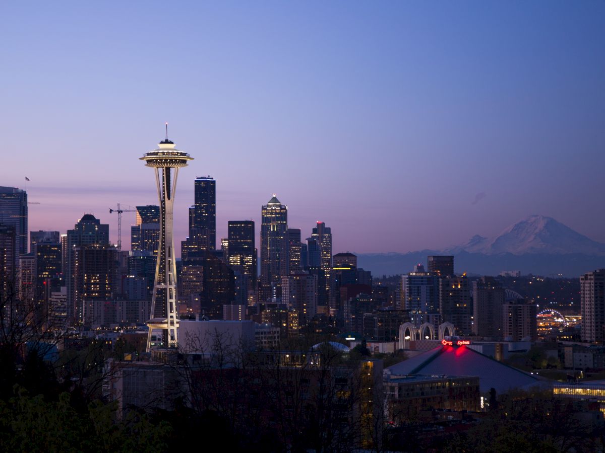 A twilight view of a city skyline featuring a prominent tower, with mountains in the background and a mix of buildings illuminated.
