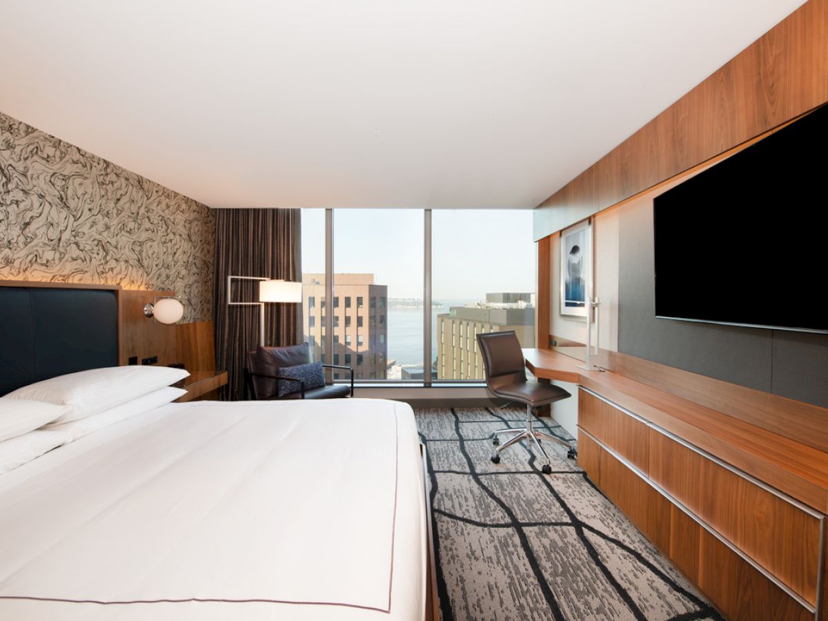 This image shows a modern hotel room with a large bed, a TV, a desk, and a city view from the window.