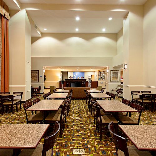 The image features an empty dining area with several tables and chairs, facing a small counter or buffet area at the far end of the room.