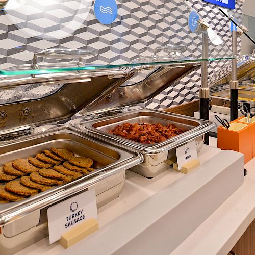 The image shows a buffet setup with trays of turkey sausage, bacon, scrambled eggs, and other breakfast items under a glass sneeze guard.