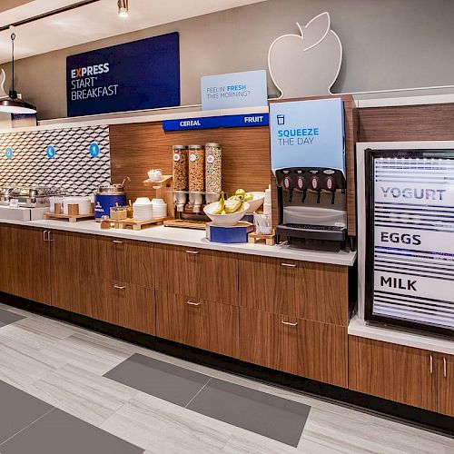 The image shows a hotel breakfast buffet setup with cereal, fruit, yogurt, eggs, and milk, along with a juice machine labeled "Squeeze The Day."