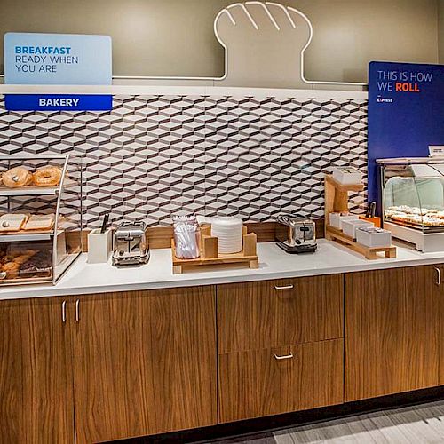 A breakfast station with pastries, bagels, toasters, and a waffle maker on a wooden counter with signs and displays.