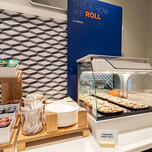 A food display area with plates, utensils, and a variety of cinnamon rolls under a sneeze guard, with a sign reading 