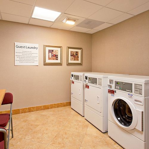 A laundry room with washers, dryers, a table, chairs, framed art, and a Guest Laundry sign on the wall, under bright lighting.