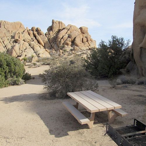 The image shows a picnic table and grill in a desert landscape with large rock formations and sparse vegetation in the background.