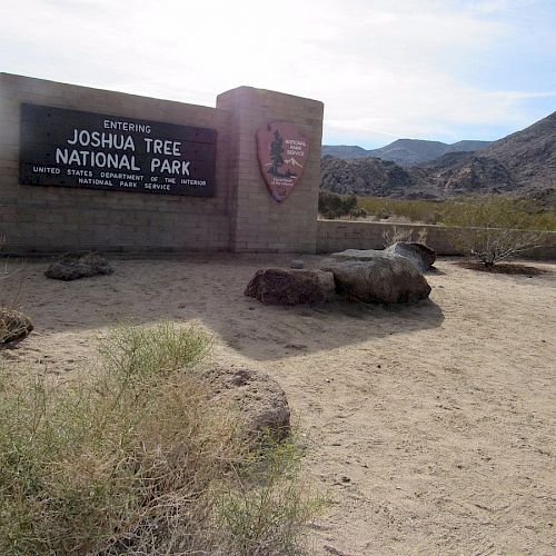 Sign at an entrance to Joshua Tree National Park, with mountains in the background and desert terrain in the foreground.