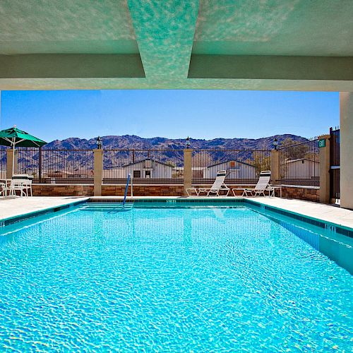 A covered swimming pool with clear blue water, featuring lounge chairs and umbrellas outside, with a mountainous landscape in the background.