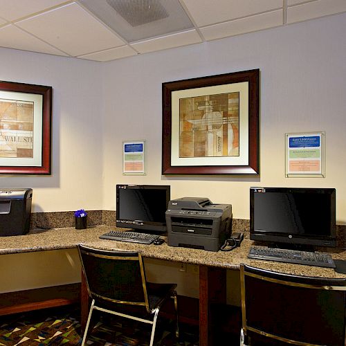 The image shows a small computer area with two desktop computers, a printer, and two chairs. The walls have framed artwork and informational signs.