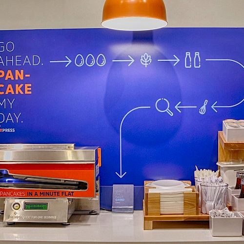 The image shows a pancake-making station with a machine, utensils, and condiments. The background instructions read 