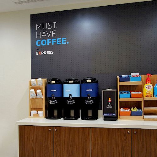 The image shows a coffee station with various coffee dispensers, cups, creamers, and sugar packets, accompanied by a "Must Have Coffee" sign.