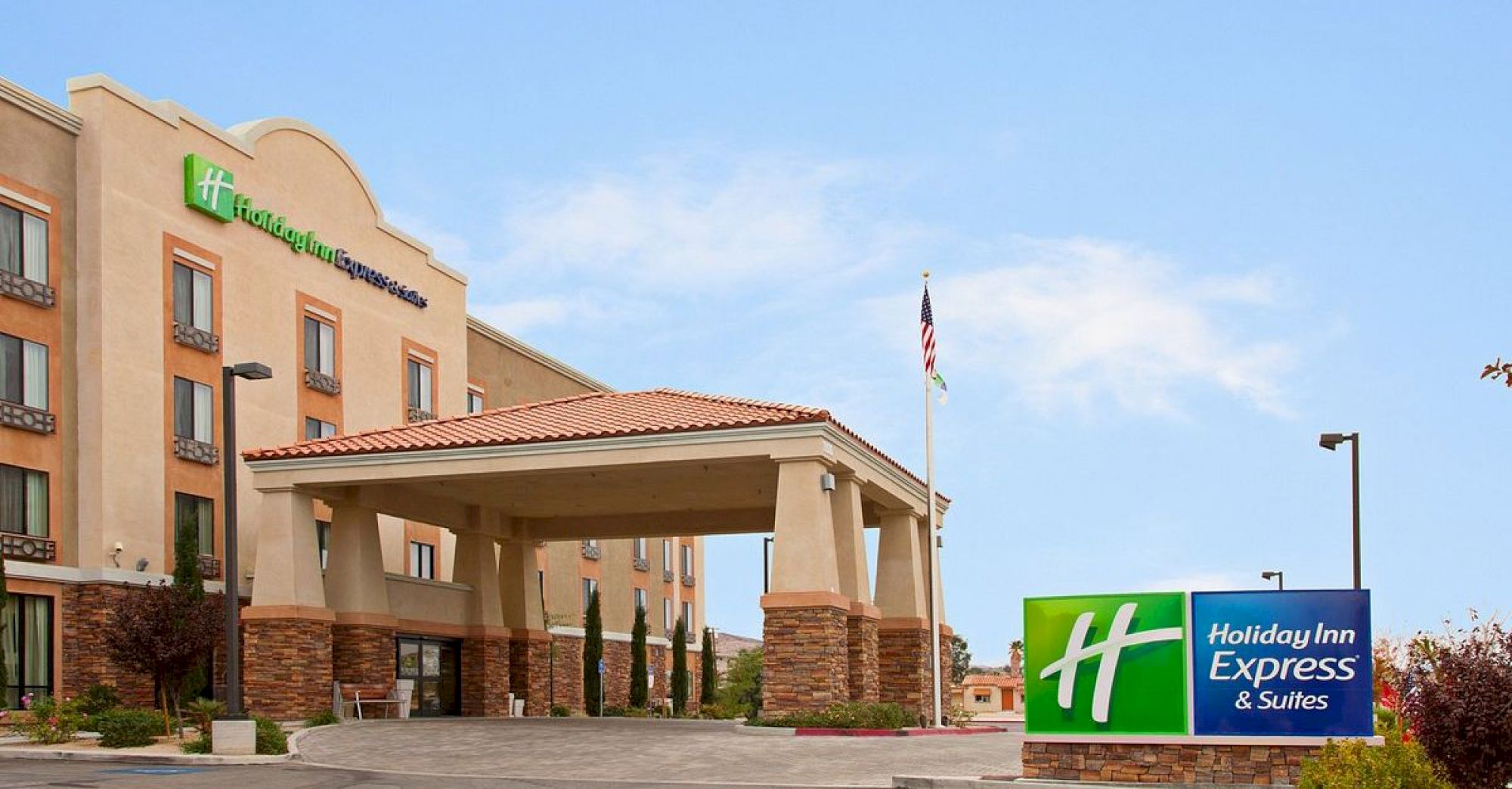 This image shows a Holiday Inn Express & Suites hotel with a prominent entrance canopy and sign in the foreground. The sky is clear blue.