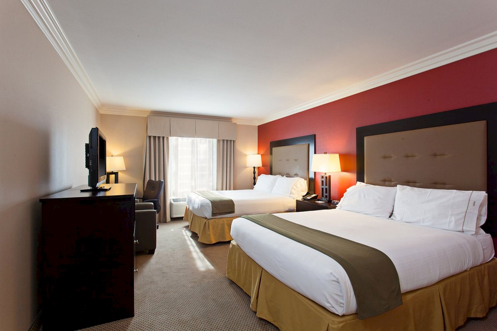 A hotel room with two beds, a TV, lamps, and a window with curtains. The wall behind the beds is red, and the room has a modern, clean look.