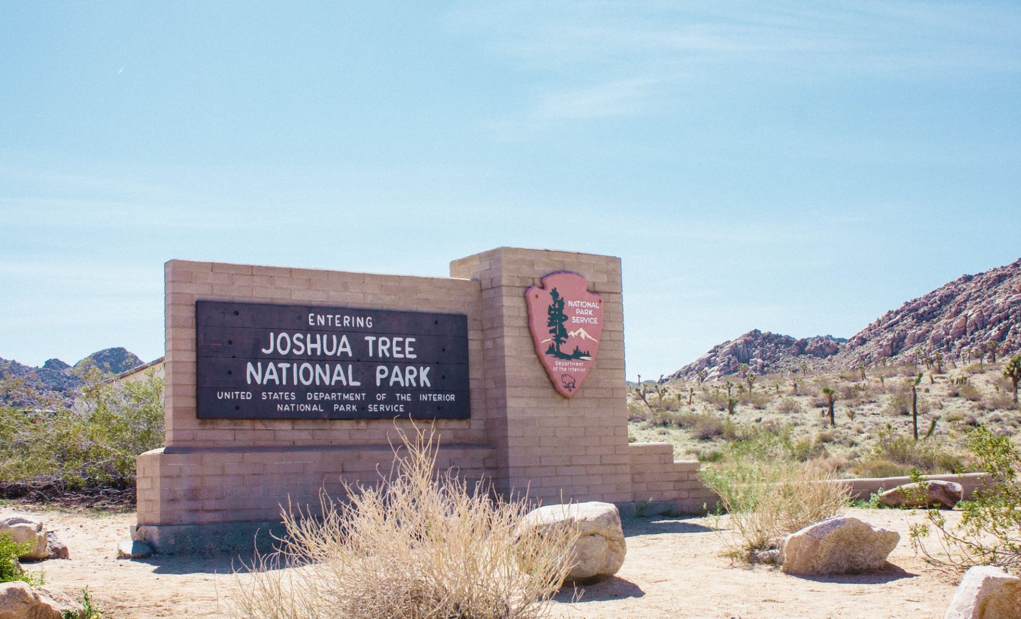 A sign at the entrance of Joshua Tree National Park with desert vegetation and rocks in the background, under a clear sky.