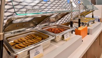 The image shows a buffet with trays of turkey sausage, cooked bacon, and other food items on a countertop, under a glass sneeze guard.