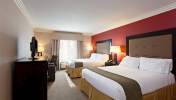 A hotel room with two double beds, a TV, a window with curtains, two lamps, and a desk chair. The walls are painted red and white, and the floor is carpeted.