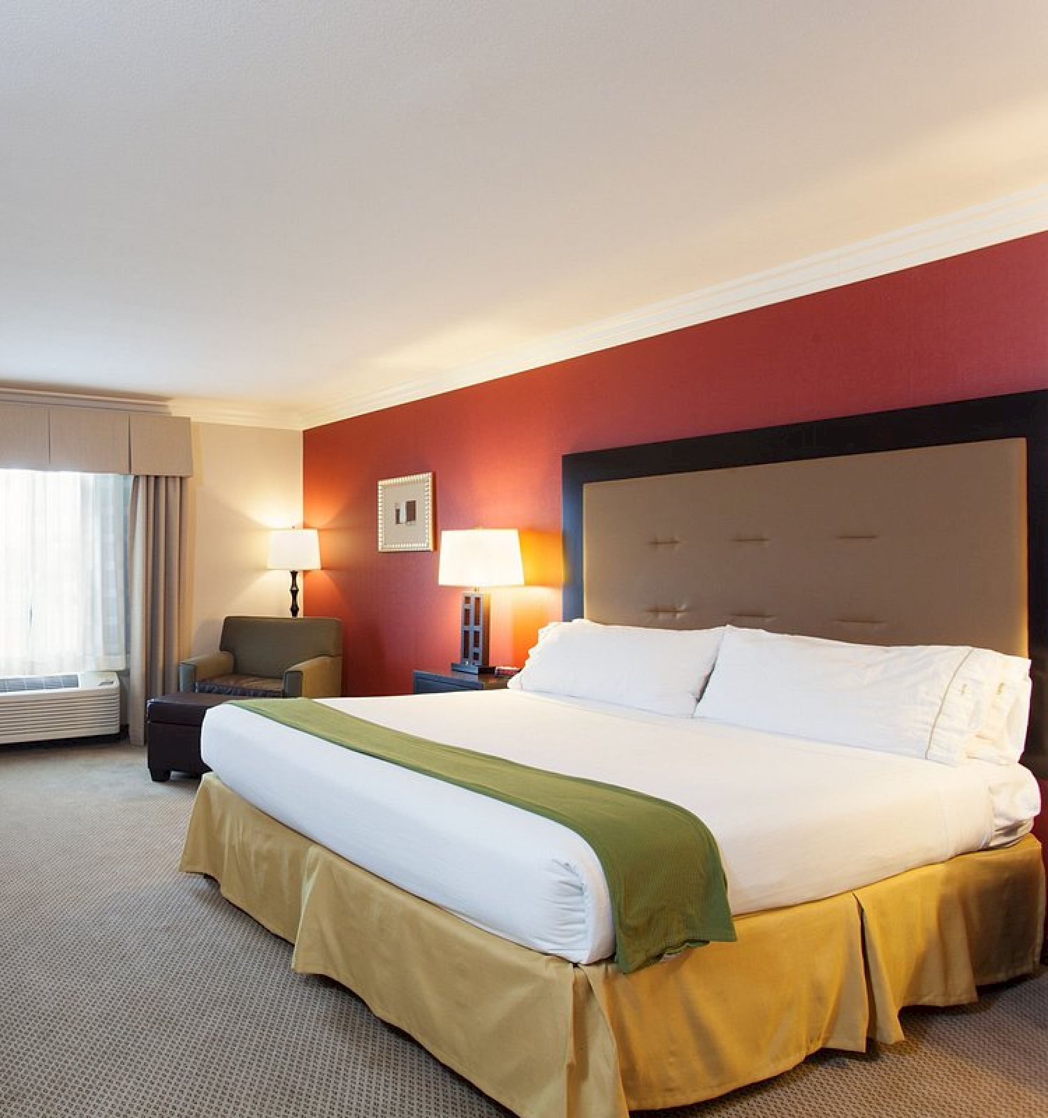 This image shows a hotel room with a large bed, TV, desk, armchair, and two lamps. The walls are red and white, and the room feels inviting.