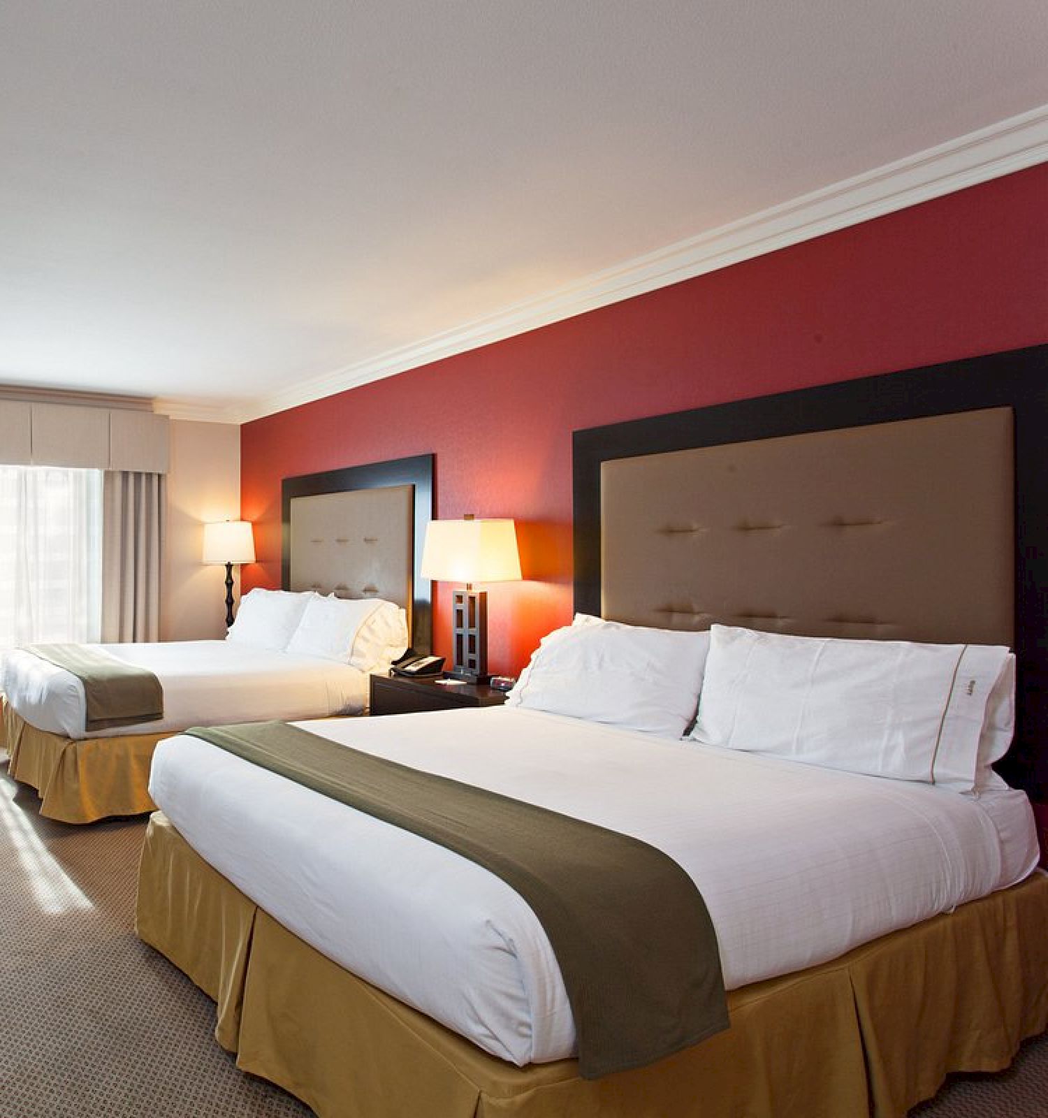 A hotel room with two double beds, a red accent wall, a dresser with a TV, a window with curtains, bedside tables with lamps, and carpeted flooring.