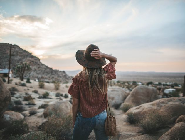 A person with long hair, wearing a hat and a brown top, stands on rocky terrain with a scenic view of mountains and a vast sky in the background.